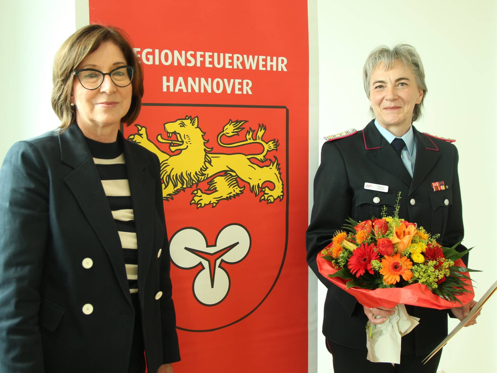 Zwei Damen vor einem großen Banner auf dem Regionsfeuerwehr Hannover steht. Eine der Damen hält einen Blumenstrauß in den Händen